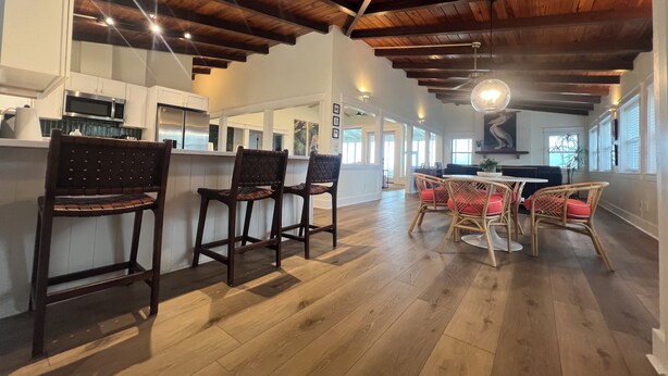 A spacious kitchen with white cabinets and a wooden ceiling. There are bar stools and a dining table with chairs in the dining area. - SATSUMA SUNSET☀️ Spacious '70s beach bungalow 🏖️ beachfront 🌊 tranquil retreat (Alligator Point)