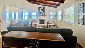 A cozy living room with a wooden ceiling, a fireplace, and a large window. - SATSUMA SUNSET☀️ Spacious '70s beach bungalow 🏖️ beachfront 🌊 tranquil retreat (Alligator Point)