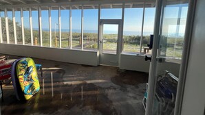 A room with a large window overlooking a beach, a door, and a surfboard. - SATSUMA SUNSET☀️ Spacious '70s beach bungalow 🏖️ beachfront 🌊 tranquil retreat (Alligator Point)