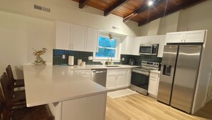 A modern kitchen with white cabinetry, a wooden ceiling, and a large island with seating. - SATSUMA SUNSET☀️ Spacious '70s beach bungalow 🏖️ beachfront 🌊 tranquil retreat (Alligator Point)