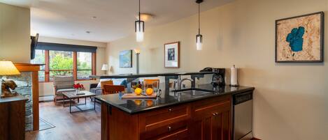 A modern kitchen with a large island, wooden cabinets, and a stone backsplash. There is a dining area with a table and chairs, and a large window providing natural light.