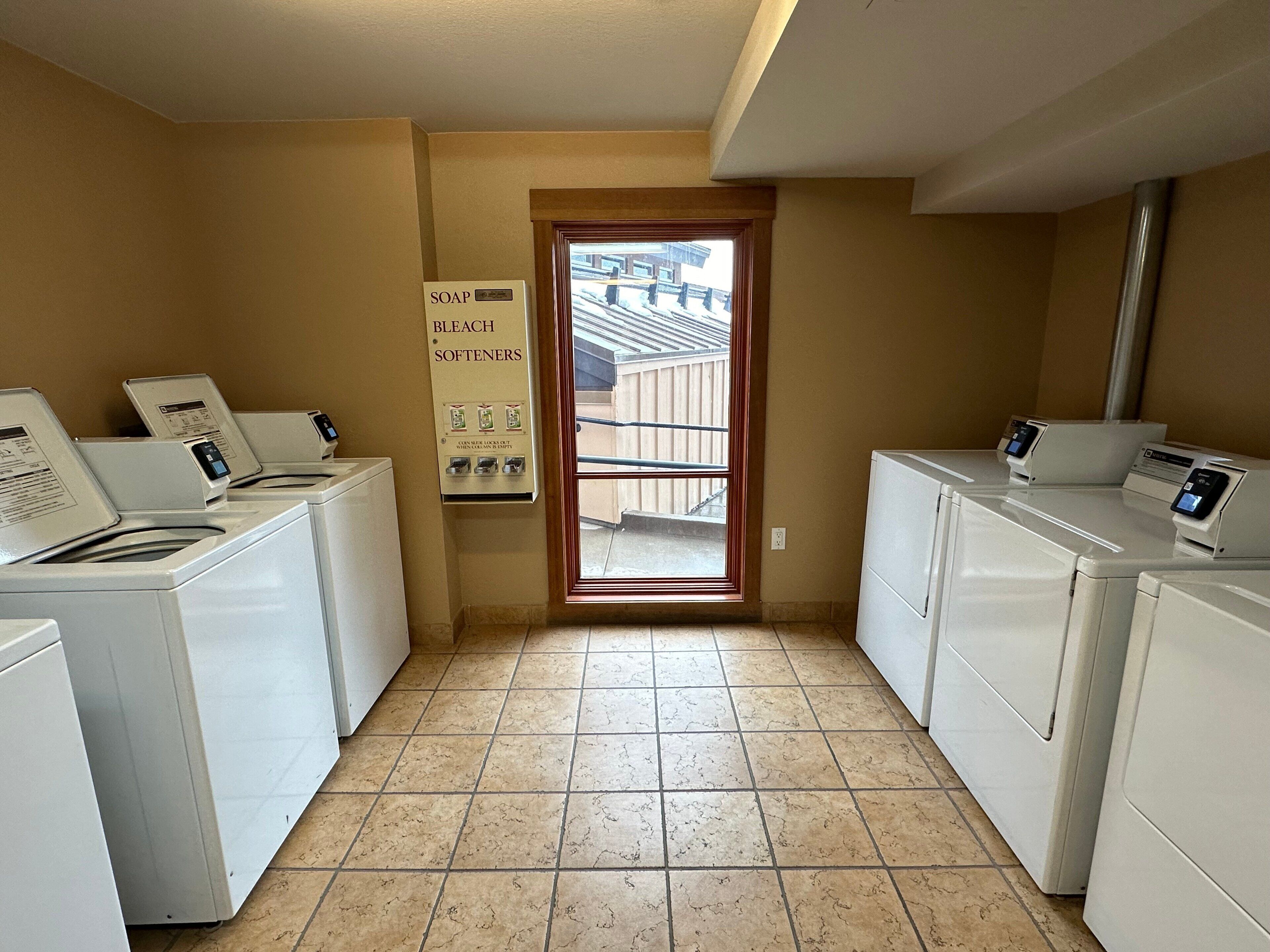 A laundry room with four washers and dryers, a sign indicating soap, bleach, and softeners, and a view of the outside through a door.