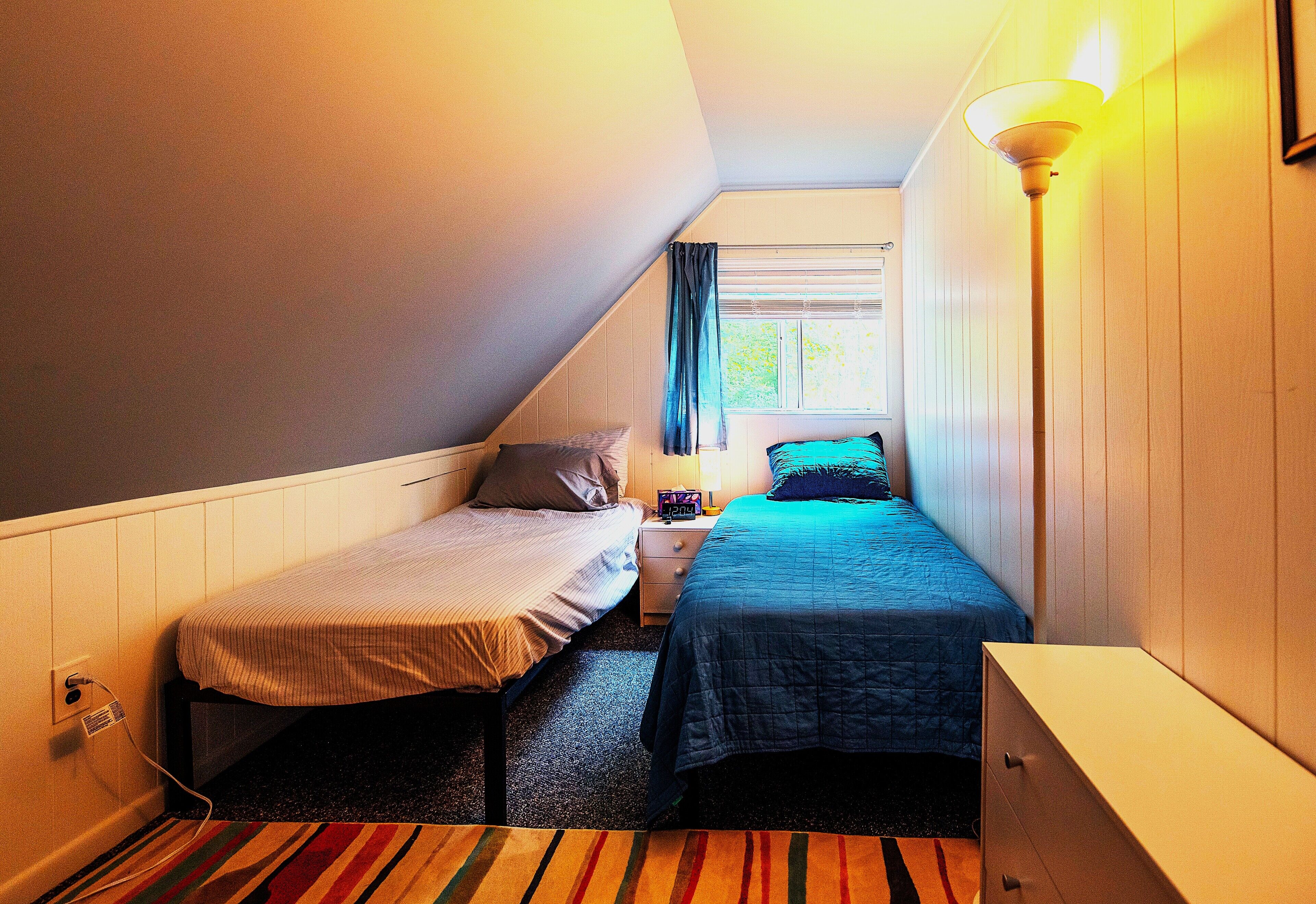 A small, narrow room with two single beds, a nightstand, a dresser, and a window with curtains.
