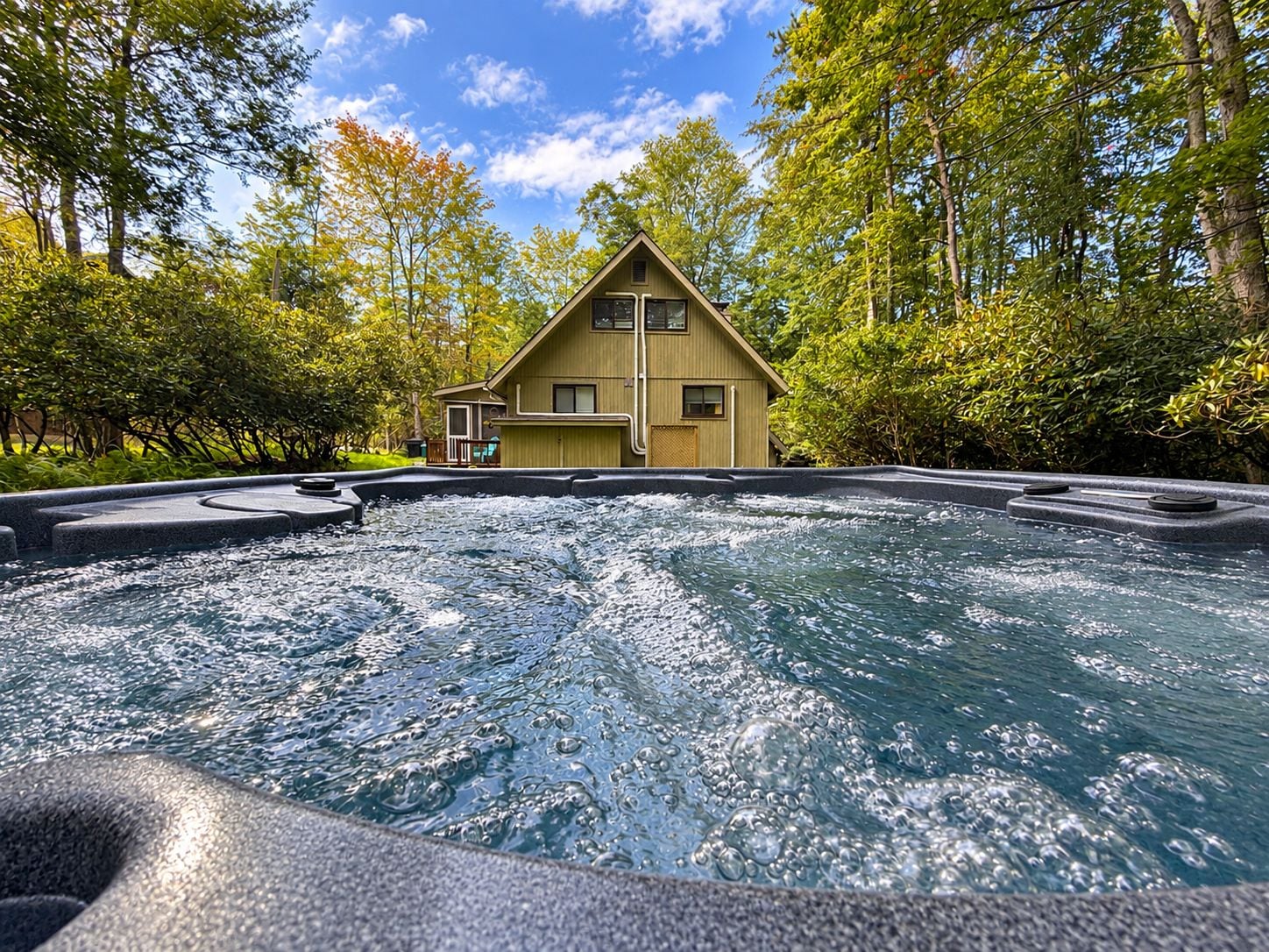 Un bain à remous avec une maison en bois en arrière-plan, le tout entouré d’arbres.