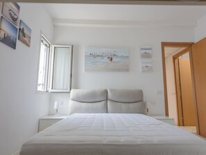 A bedroom with a bed, a nightstand, a window with curtains, a painting on the wall, and a door.