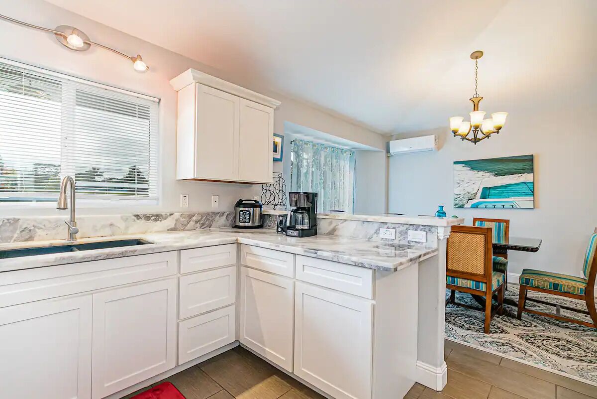 A kitchen with white cabinets, a marble countertop, a coffee maker, and a dining area with chairs and a rug.