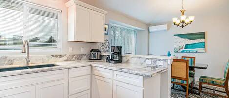 A kitchen with white cabinets, a marble countertop, a coffee maker, and a dining area with chairs and a rug.