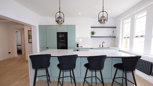 A modern kitchen with a central island, three black bar stools, and pendant lighting. - Newly Refurbished w/ River Views (Greater London)