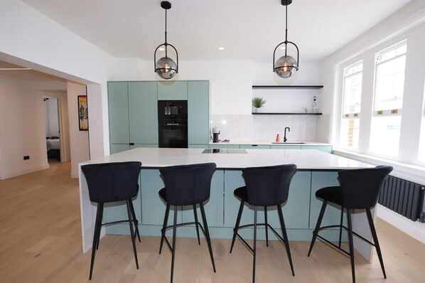 A modern kitchen with a central island, three black bar stools, and pendant lighting. - Newly Refurbished w/ River Views (Greater London)