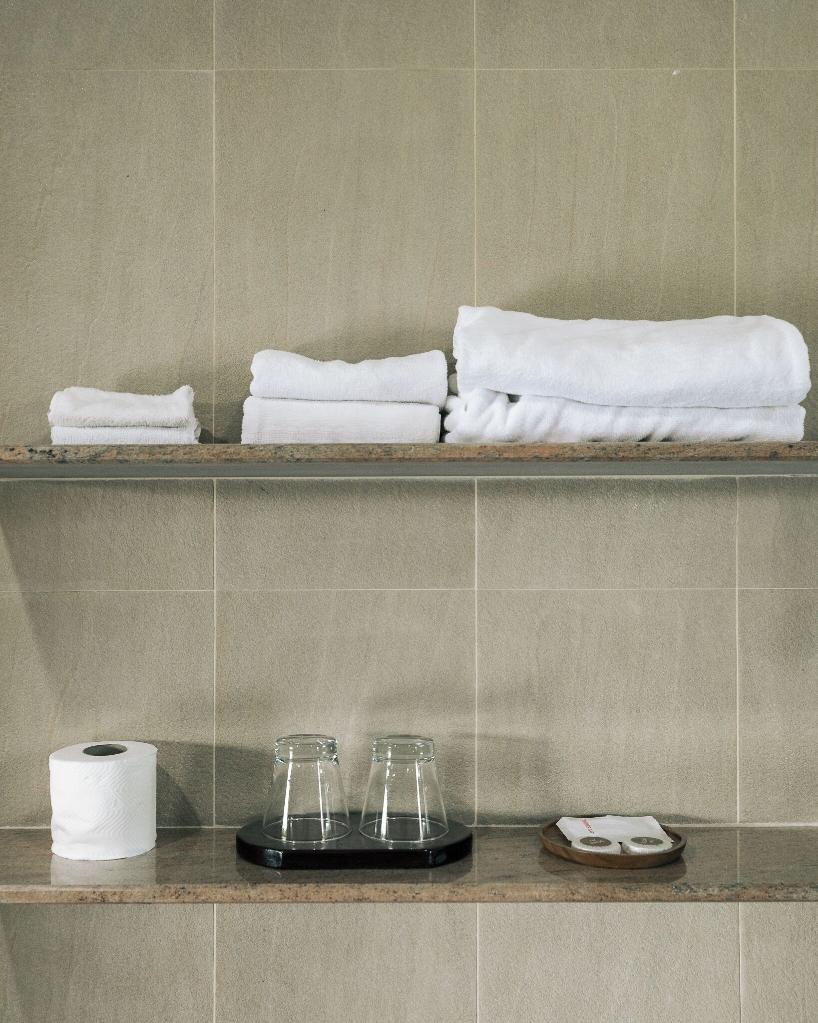 Une salle de bain avec un porte-serviettes où sont pliés des serviettes, un rouleau de papier toilette et un savonnière contenant deux flacons.