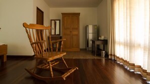A wooden rocking chair in a room with a wooden floor, a kitchen area with a refrigerator, and a window with curtains. - Thaihome Resort (Bang Kruai)