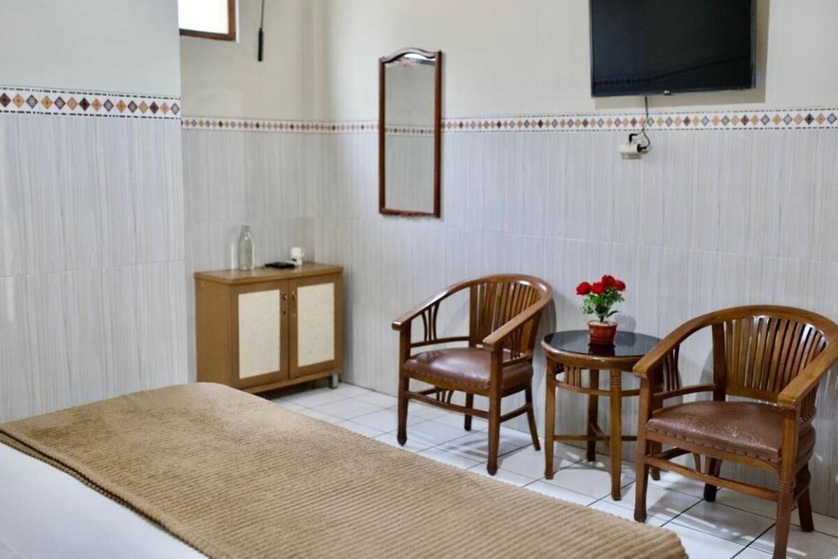 A hotel room with a bed, two wooden chairs, a small table with a potted plant, a wooden cabinet, and a TV mounted on the wall.