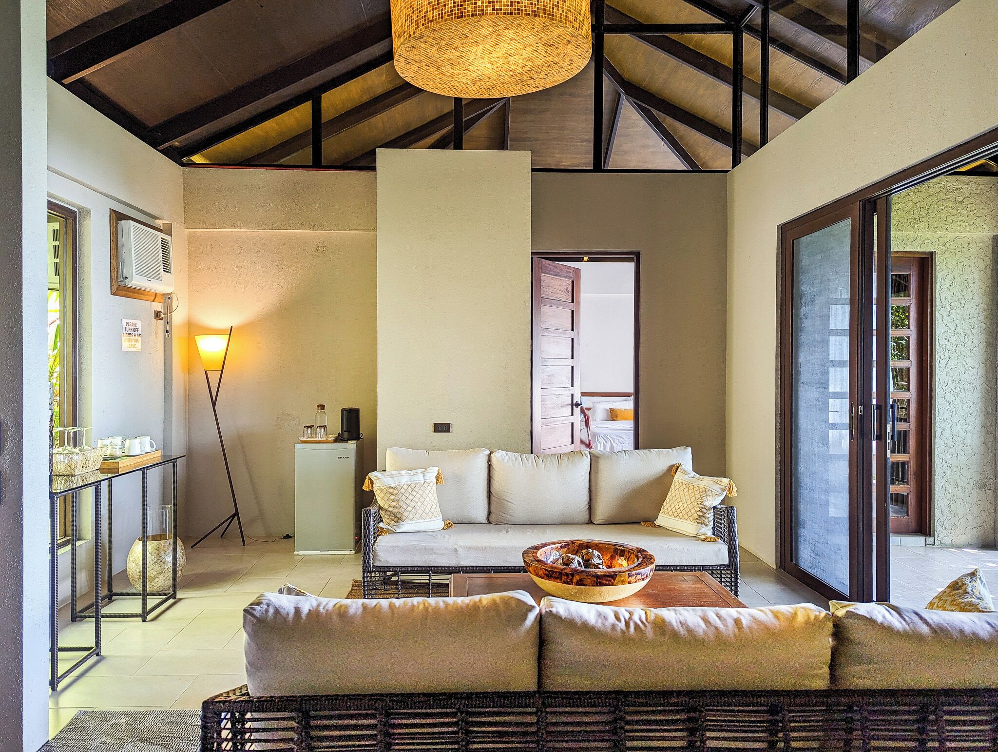 A spacious living area with a sofa set, a coffee table, and a dining table. There is a large window providing natural light and a view of the outdoors.