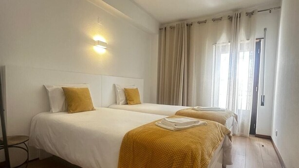 A hotel room with two beds, a bedside table, a lamp, and a window with curtains. - João Paulo II - Fátima (Fátima)