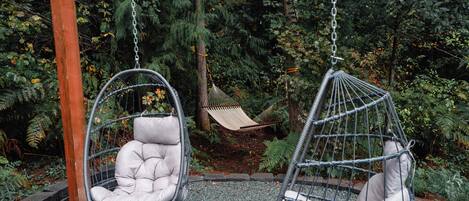 Zwei Hängestühle in einer Gartenumgebung mit einer Hängematte und üppiger Bepflanzung im Hintergrund.