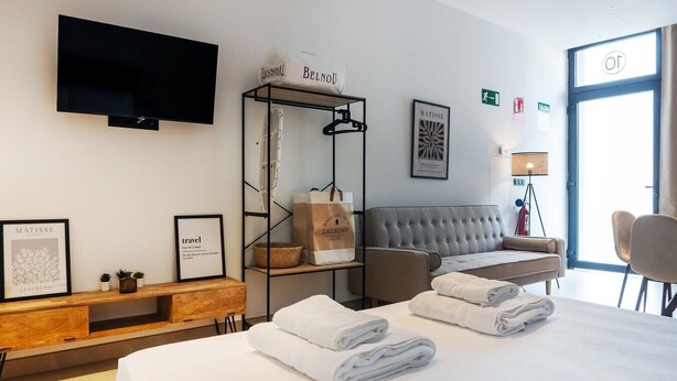 A hotel room with a bed, a grey sofa, a wooden console table, a TV mounted on the wall, and a large window. - VLC HOST - Zapadores J (Valencia)