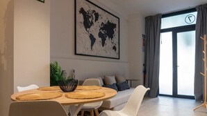 A modern living room with a round wooden table, white chairs, a sofa with cushions, a world map artwork on the wall, and a large window with curtains.