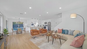 Un amplio salón con sofá, sillas y una mesa de centro. Se ve una cocina con armarios blancos y un comedor con sillas al fondo.