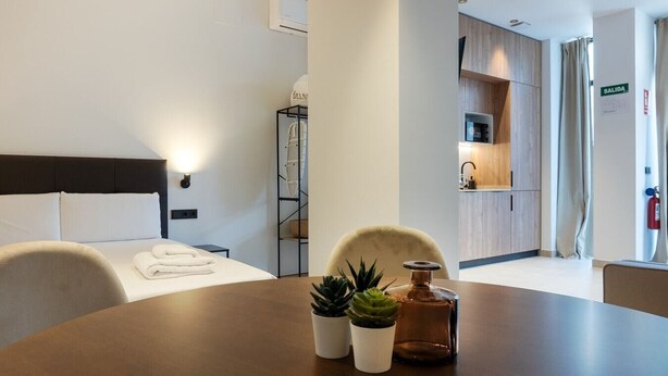 A modern hotel room with a bed, a dining table with potted plants, a kitchen area, and a visible exit sign. - VLC HOST - Zapadores I (Valencia)