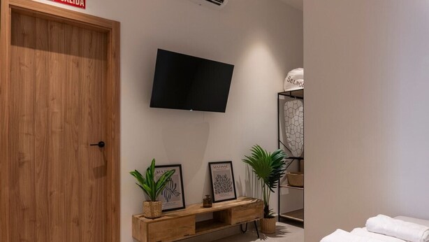 A hotel room with a wooden door, a flat-screen TV mounted on the wall, a wooden console table with two framed artworks, a potted plant, and a neatly folded white towel on the floor. - VLC HOST - Zapadores E (Valencia)