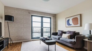 Ett modernt vardagsrum med en grå soffa, ett soffbord, en golvlampa och ett stort fönster med utsikt över en byggnad.