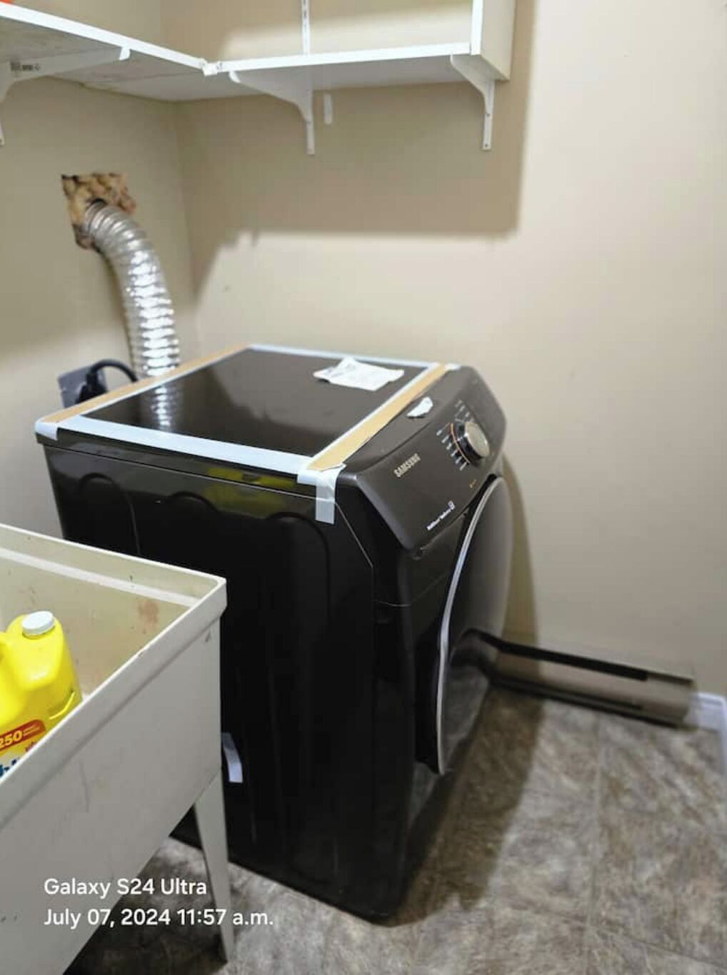 A compact laundry room with a front-loading washer and dryer, a small storage bin, and a visible ductwork on the wall.