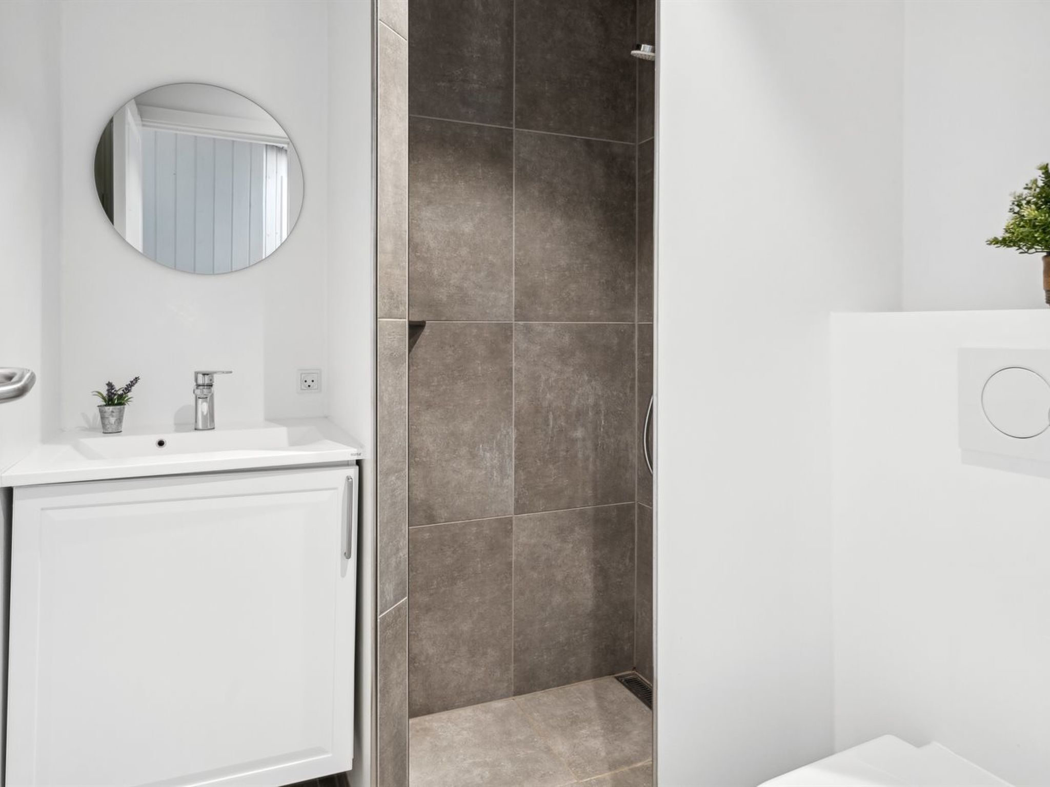 Un baño moderno con un tocador blanco, un espejo redondo, una pequeña planta en maceta y una zona de ducha con azulejos grises.