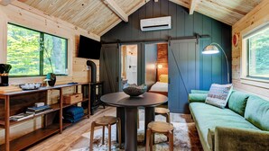 A cozy cabin interior with a wooden ceiling, a round table with stools, a green sofa, and a large window offering a view of the outdoors. - 2 Bedroom Cabin in DuPont State Park with Loft, Hot-Tub, and Sauna(Read Rules) (Hendersonville)