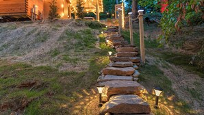 A stone pathway leading to a wooden cabin with string lights, surrounded by greenery. - 2 Bedroom Cabin in DuPont State Park with Loft, Hot-Tub, and Sauna(Read Rules) (Hendersonville)