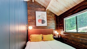 A wooden cabin bedroom with a bed, a framed picture on the wall, and a window with a view of the forest. - 2 Bedroom Cabin in DuPont State Park with Loft, Hot-Tub, and Sauna(Read Rules) (Hendersonville)
