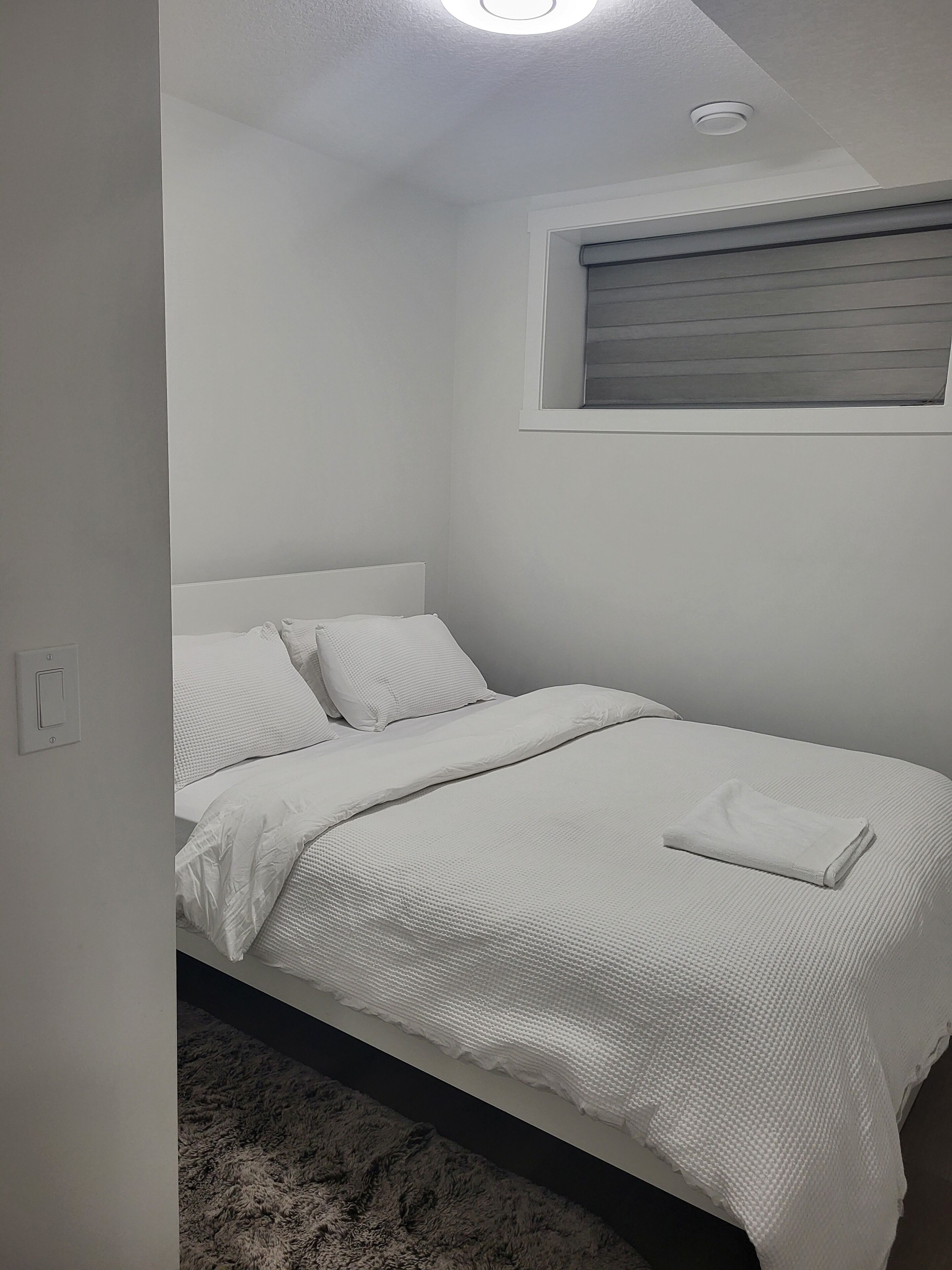 A neatly made bed with white bedding in a room with white walls and a window with blinds.