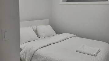A neatly made bed with white bedding in a room with white walls and a window with blinds.