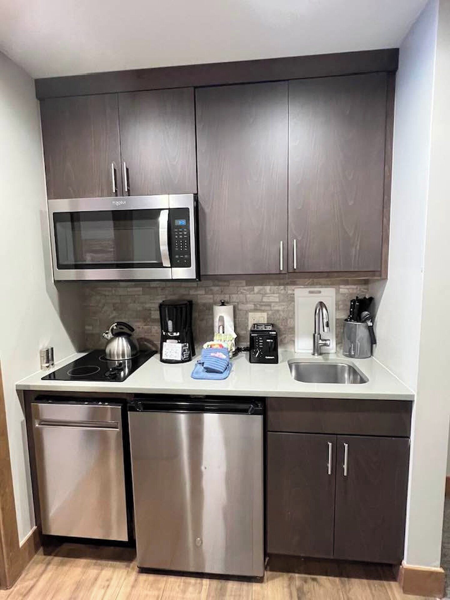 A compact kitchen with dark cabinets, a stainless steel microwave, a coffee maker, a kettle, and a dishwasher.