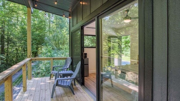 Une terrasse extérieure couverte avec un plancher en bois, deux fauteuils Adirondack et une porte vitrée donnant sur un espace patio.