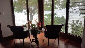 Een kamer met een houten vloer, twee stoelen, een tafel met bloemen en een groot raam met uitzicht op water en groen.