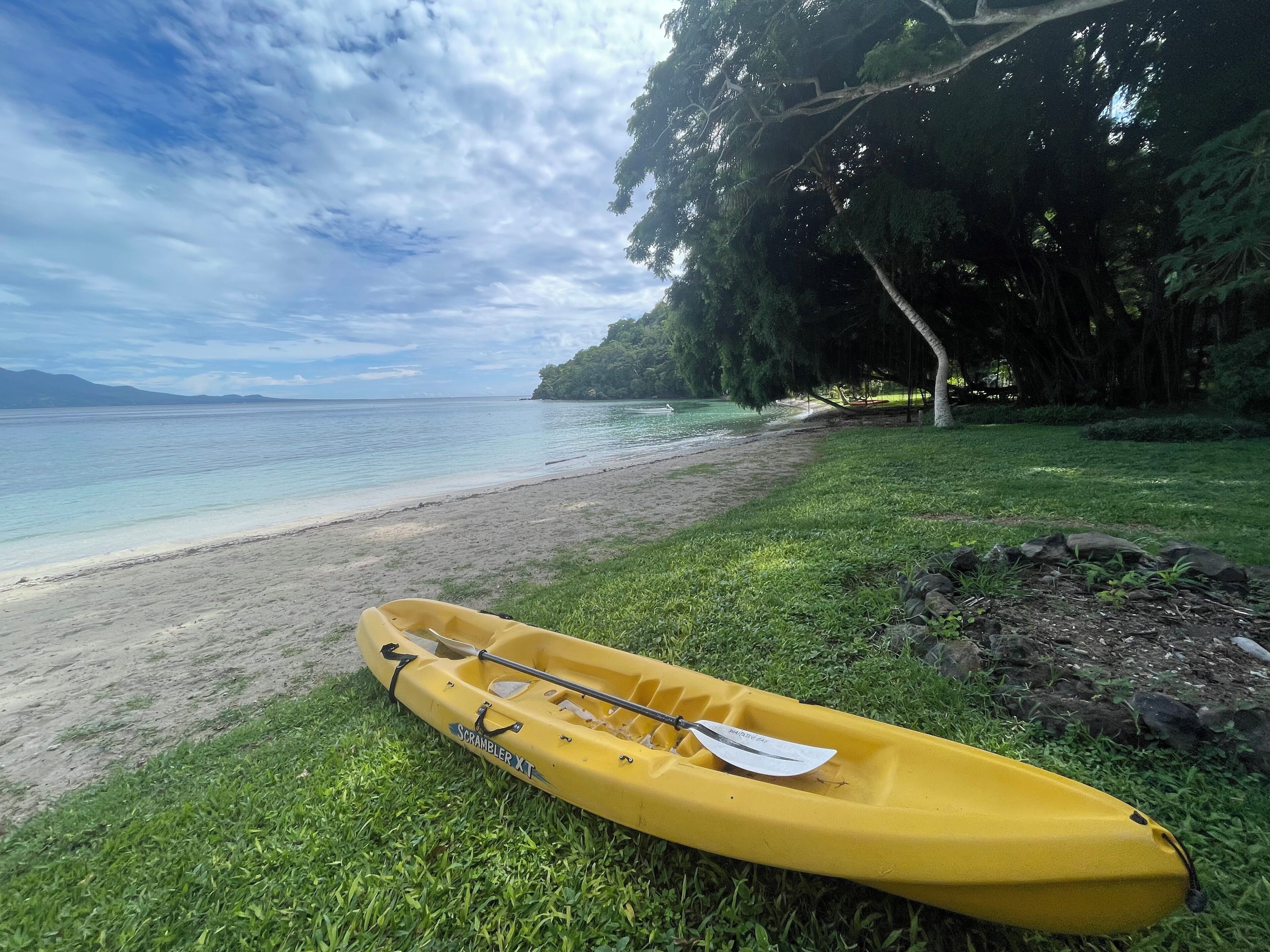 A yellow kayak on a grassy area near a beach with clear water and a tree line in the background.