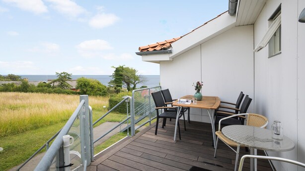 A balcony with a wooden table and chairs, overlooking a grassy area and the sea. - Gudhjem Søpark Lejl. 24 (Gudhjem)