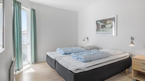 A bedroom with a bed, two bedside lamps, a window with curtains, a bedside table, and a framed picture on the wall. - Gudhjem Søpark Lejl. 24 (Gudhjem)