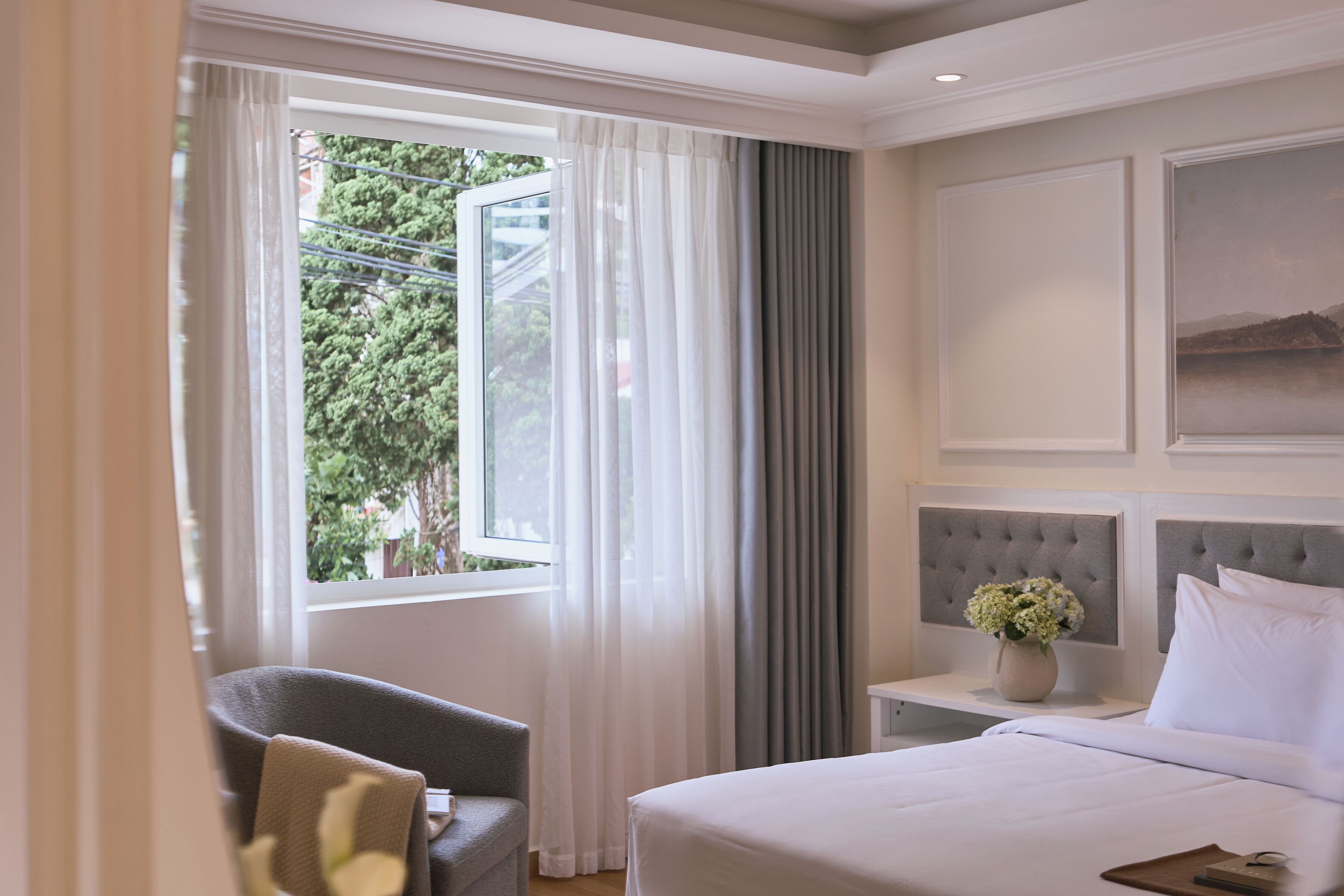 Una camera d'hotel con un letto, una poltrona, un comodino con un vaso di fiori e una finestra con tende.