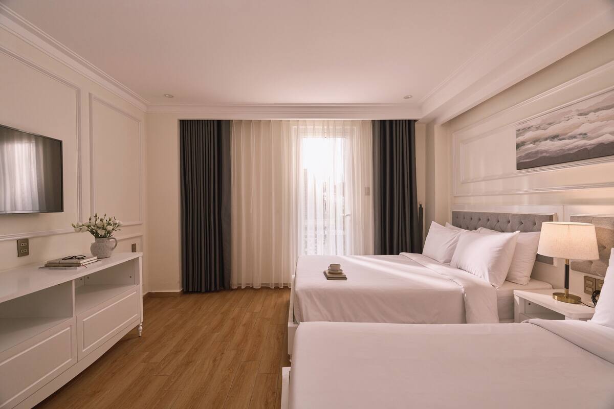 Habitación de hotel con dos camas, televisión, mesita de noche con lámpara, un cuadro en la pared y una ventana con cortinas.