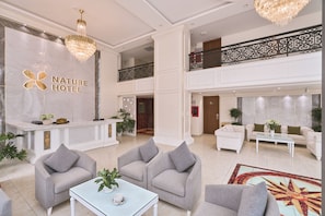 A hotel lobby with a reception desk, grey sofas, a chandelier, and a decorative railing. - Nature Hotel - Pham Hong Thai (Da Lat)