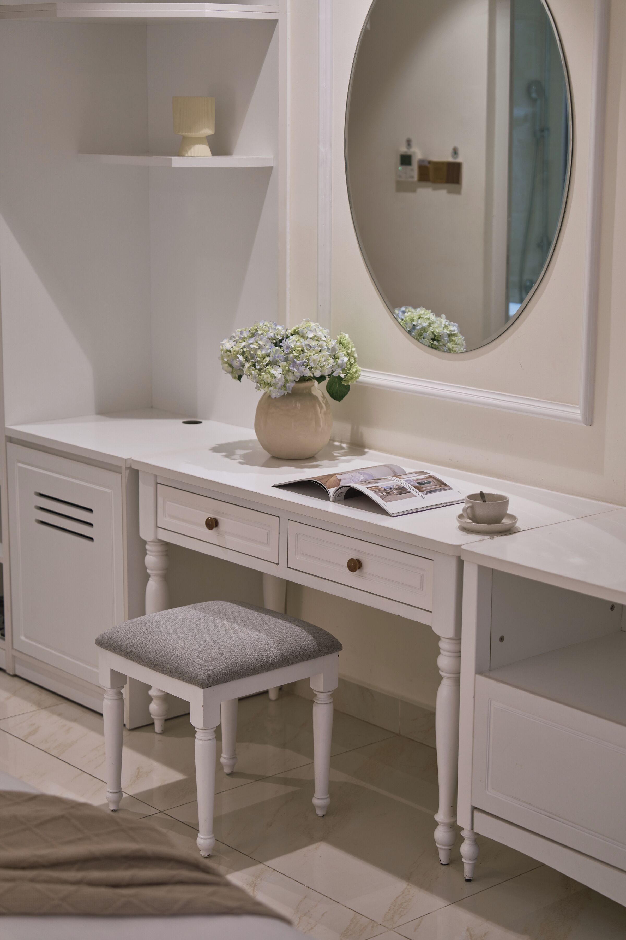 Une coiffeuse blanche avec un miroir, un vase de fleurs, un magazine et une tasse.