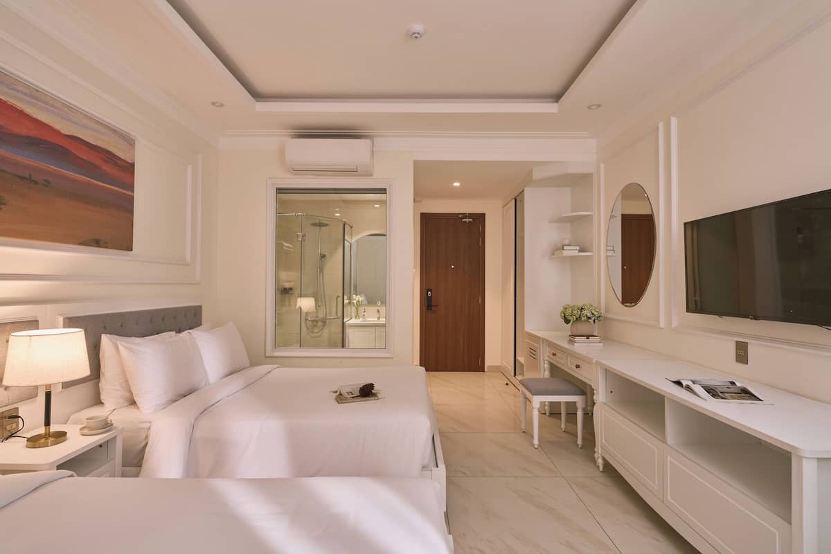 Habitación de hotel con dos camas, un escritorio, televisor y baño con ducha de vidrio.