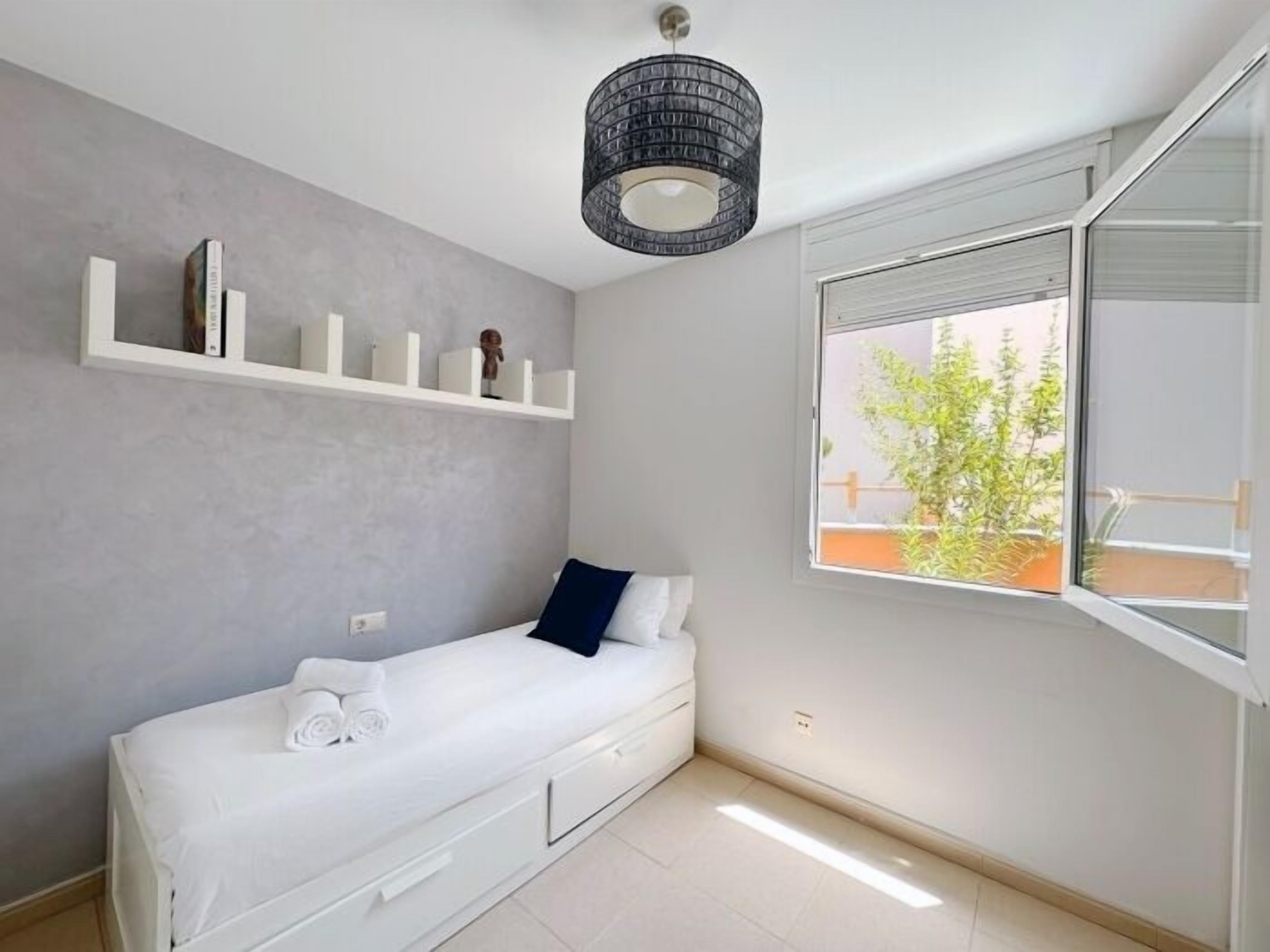 Un dormitorio minimalista con una cama individual, una ventana que da a vistas de vegetación y una estantería con libros y una pequeña figurita.