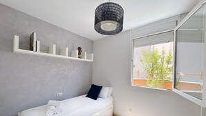 Et minimalistisk soveværelse med en enkelt seng, et vindue med udsigt til grøn natur, og en reol med bøger og en lille figur.