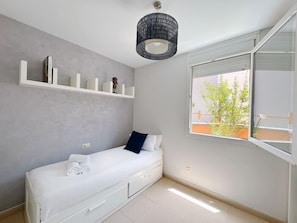 A minimalist bedroom with a single bed, a window with a view of greenery, and a shelf with books and a small figurine. - Apartment with large terrace in Costa Adeje. (Costa Adeje)