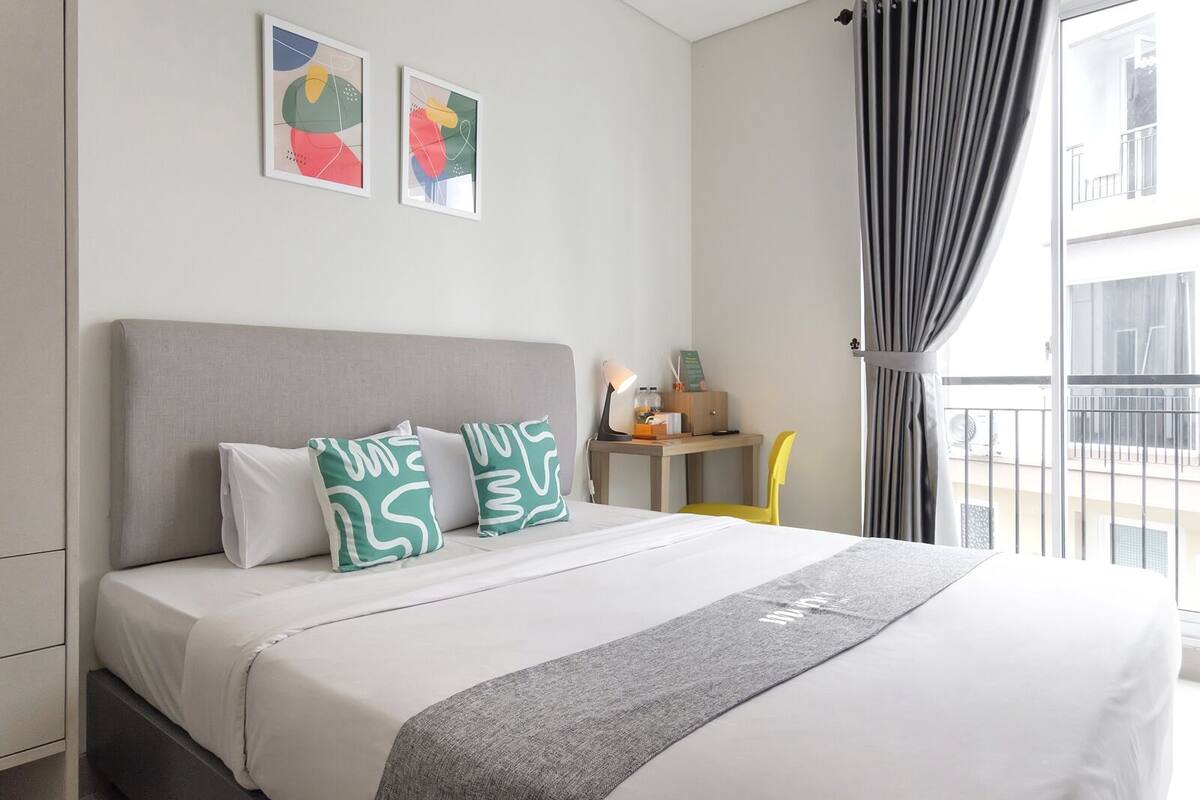 A neatly made bed with a grey headboard, two pillows with a green and white abstract pattern, and a grey throw blanket. There are two framed abstract paintings on the wall above the bed.