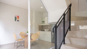 A modern kitchen with white cabinets, a dining area with a glass partition, and a black metal staircase. - Mono Coliving at BSD City (Pagedangan)