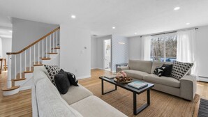 A modern living room with a staircase, a sofa set, a coffee table, and a view through a large window. - Stunning Cape Home Near Chatham (Brewster)