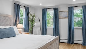 A bedroom with a bed, a nightstand, a lamp, a plant, and a window with curtains. - Stunning Cape Home Near Chatham (Brewster)